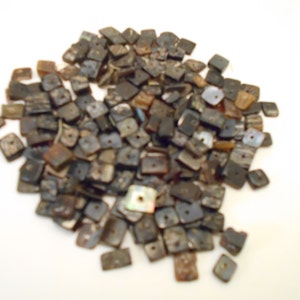 May include: A pile of small, square beads with a hole in the center. The beads are primarily dark brown and black, with some lighter, iridescent patches. The beads are scattered on a white surface.