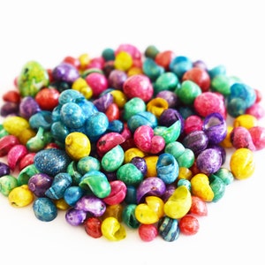 May include: A large pile of small, polished sea shells dyed in a vibrant array of colors including bright yellow, hot pink, deep purple, turquoise, lime green, and red. Each shell features a smooth, glossy finish and a distinct spiral shape.