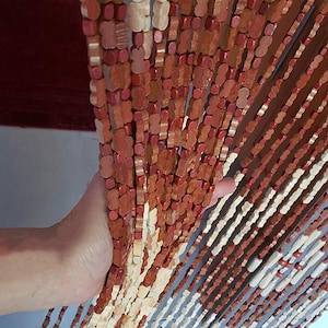 May include: A wooden beaded curtain with alternating light brown and reddish-brown beads. The curtain is hanging from a doorway and is being held open by a hand.