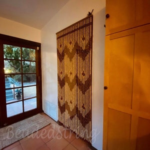 May include: A beaded door curtain with a geometric pattern in shades of brown, tan, and olive green hangs on a white wall. The curtain is made of wooden beads and is suspended from a dark brown wooden header. The words "BeadedString" are visible at the bottom.