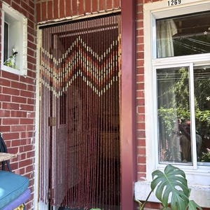 May include: A beaded door curtain featuring a chevron pattern in shades of brown, cream, and orange. The curtain consists of numerous vertical strands of small beads suspended from a horizontal rod, creating a semi-transparent room divider.