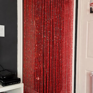 K&ouml;nnte beinhalten: Ein roter Perlenvorhang h&auml;ngt in einem T&uuml;rrahmen. Der Vorhang besteht aus vielen Str&auml;ngen kleiner, rechteckiger Perlen. Die Perlen haben eine leuchtend rote Farbe. Der T&uuml;rrahmen ist wei&szlig; und die Wand links ist dunkelgrau.