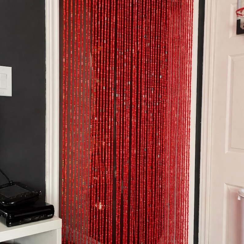 K&ouml;nnte beinhalten: Ein roter Perlenvorhang h&auml;ngt in einem T&uuml;rrahmen. Der Vorhang besteht aus vielen Str&auml;ngen kleiner, rechteckiger Perlen. Die Perlen haben eine leuchtend rote Farbe. Der T&uuml;rrahmen ist wei&szlig; und die Wand links ist dunkelgrau.