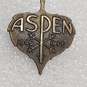 May include: A bronze-colored pin shaped like an aspen leaf with the word "ASPEN" in raised letters and the years "1946" and "1966" on either side of a stylized snowflake.