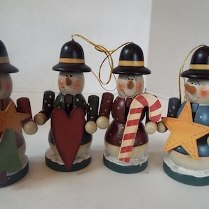 May include: Set of four wooden snowman ornaments with different decorations. Each snowman is wearing a black hat with a yellow band and has a painted face. The ornaments are holding a star, a heart, a candy cane, and a Christmas tree.