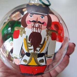 May include: A glass Christmas ornament featuring a hand-painted nutcracker. The nutcracker is wearing a white uniform with red and gold accents. The ornament is clear and has a green wire hanger.