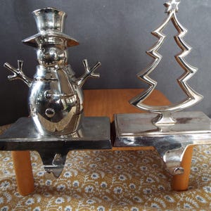 May include: Two silver Christmas stocking holders. One is a snowman with a top hat and scarf, the other is a Christmas tree with a star on top. Both have a rectangular base and a wooden peg.
