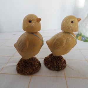 May include: Two wooden chick figurines with yellow bodies, brown bases, and small orange beaks. Each chick has a light blue ribbon around its neck. The figurines are decorated with a speckled finish, giving them a rustic appearance.