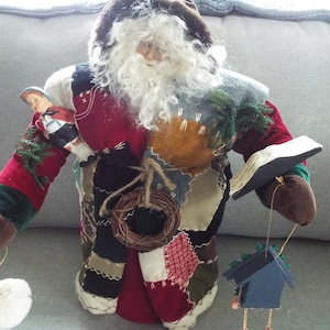 May include: A patchwork Santa Claus doll with a white beard and a book in one hand. The doll is wearing a red, green, and blue patchwork outfit and has a small doll in its other arm. The doll is holding a wreath and a birdhouse.