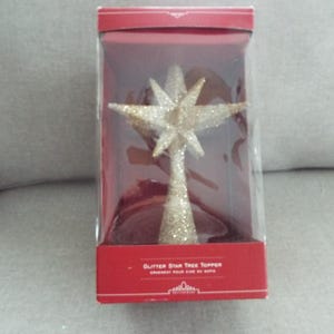 May include: A gold glitter star tree topper in a clear plastic box with a red base and lid. The box has the text "Glitter Star Tree Topper" on the front. The star is a festive holiday decoration.