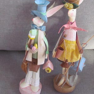 May include: Two Easter bunny figurines, one wearing a blue top hat and holding a basket of eggs, the other wearing a yellow skirt and holding gardening tools.