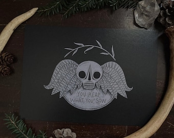 May include: A black print featuring a skull with feathered wings and the text "YOU REAP WHAT YOU SOW" in a circular design. The artwork is surrounded by natural elements like pine needles, pine cones, and antlers, creating a rustic aesthetic.