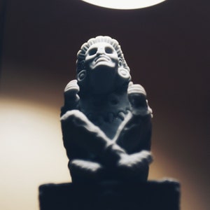 Peut inclure: Une sculpture en céramique blanche d'une personne avec les bras croisés, portant une coiffe et des bijoux. La sculpture est posée sur une base noire.