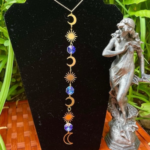 May include: A gold-colored celestial-themed necklace featuring crescent moons, sunbursts, and blue crystal beads. The necklace is displayed on a black stand, with a statue in the background. The necklace is a delicate piece of jewelry.