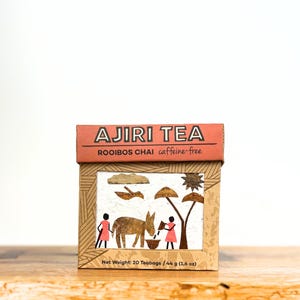 May include: A box of Ajiri Tea Rooibos Chai, caffeine-free. The box features a decorative design with a brown donkey, two figures in pink dresses, and a tree. The net weight is 20 teabags / 44 g (1.6 oz).