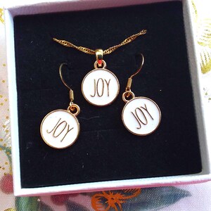 May include: A gold-toned jewelry set consisting of a pendant necklace and two matching drop earrings, all featuring circular white enamel charms with the word JOY printed in black, presented in a square white box with a black velvet insert.