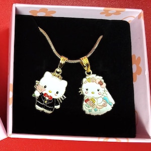 May include: A gold chain necklace with two pendants featuring cartoon bride and groom Hello Kitty characters. The bride is wearing a white dress with a blue veil and holding a bouquet of flowers. The groom is wearing a black tuxedo and holding a red rose.