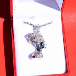 May include: A silver-toned pendant necklace featuring the profile of Queen Nefertiti. The pendant is encrusted with numerous small, clear, sparkling stones across the crown and base, suspended from a twisted rope chain, presented in a red box.