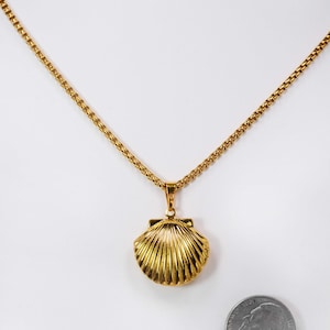 May include: A gold-colored necklace featuring a seashell pendant. The pendant has a detailed, ridged design. The necklace chain is also gold and has a delicate, linked pattern. A nickel coin is next to the necklace for size comparison.