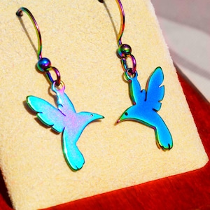 May include: A pair of iridescent hummingbird earrings. The earrings feature a vibrant color gradient of blues, greens, and purples. The hummingbird design is detailed with outstretched wings and a pointed beak. The earrings are displayed on a beige surface.
