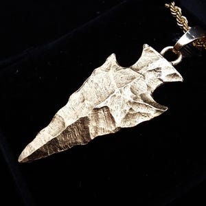 May include: A gold-toned arrowhead pendant with a textured, chipped surface finish, suspended from a matching gold-toned rope chain. The pendant features a pointed tip and notched base, reflecting light across its uneven, metallic surface.
