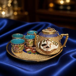 May include: A decorative tea set with a gold-colored teapot, four cups, and a matching tray. The teapot and cups have a blue interior and are decorated with floral and bird designs. The set is displayed on a blue satin surface.