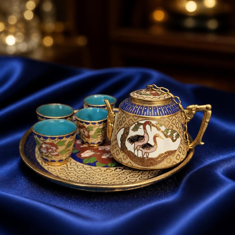 May include: A decorative tea set with a gold-colored teapot, four cups, and a matching tray. The teapot and cups have a blue interior and are decorated with floral and bird designs. The set is displayed on a blue satin surface.