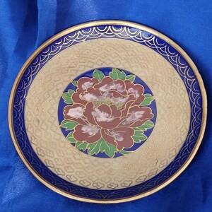 May include: A round decorative plate with a floral design. The plate features a maroon and pink peony in the center, surrounded by a blue border and gold trim. The background is beige with a textured pattern.