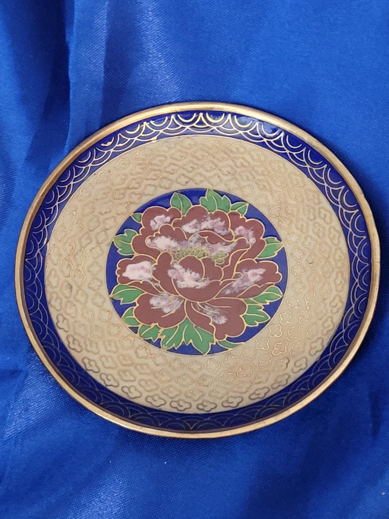 May include: A round decorative plate with a floral design. The plate features a maroon and pink peony in the center, surrounded by a blue border and gold trim. The background is beige with a textured pattern.