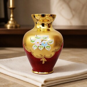 May include: A decorative vase with a gold-colored top and body, transitioning to a deep red base. The gold section features hand-painted flowers in pastel colors. The vase is likely made of glass or ceramic and is sitting on a white cloth.