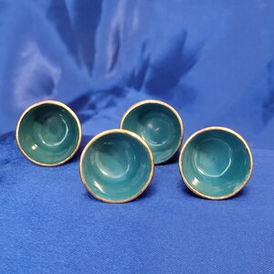 May include: Four small, teal-colored bowls with gold-colored rims are arranged on a blue surface. The bowls have a smooth, glossy finish and a simple, elegant design. The bowls are likely for serving small portions of food or for decorative purposes.