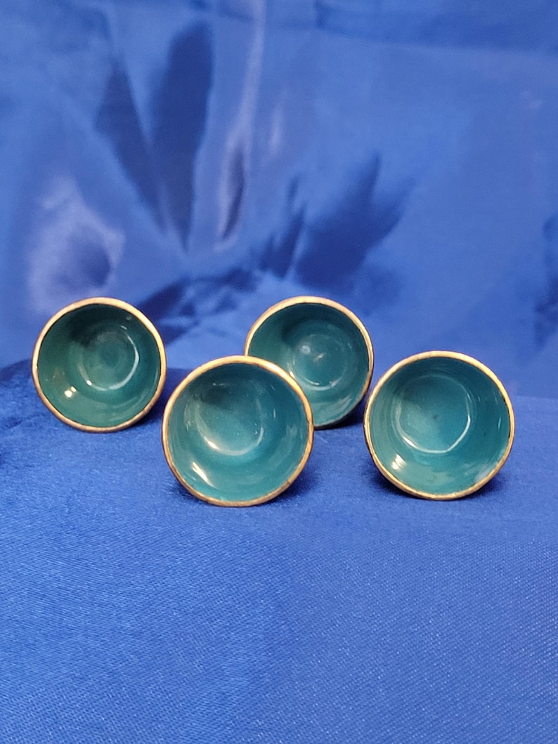 May include: Four small, teal-colored bowls with gold-colored rims are arranged on a blue surface. The bowls have a smooth, glossy finish and a simple, elegant design. The bowls are likely for serving small portions of food or for decorative purposes.