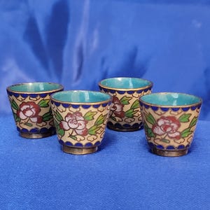 May include: Four small, decorative cups with a floral design. The cups have a beige background with red and white flowers and green leaves. The rims are blue, and the interiors are turquoise. The bases are gold-colored.