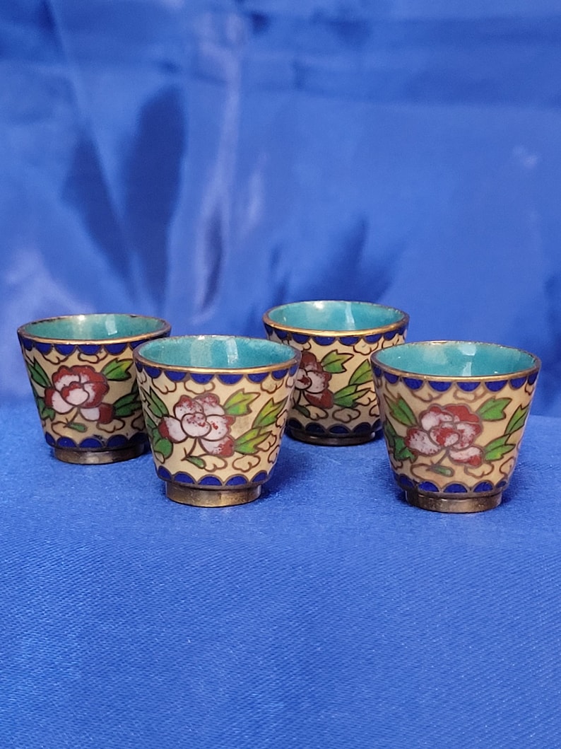 May include: Four small, decorative cups with a floral design. The cups have a beige background with red and white flowers and green leaves. The rims are blue, and the interiors are turquoise. The bases are gold-colored.