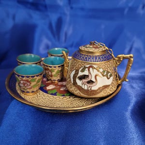 May include: Miniature tea set with a gold-colored tray, teapot, and four cups. The teapot and cups have a teal interior and are decorated with gold, blue, and red floral patterns. The teapot features a crane design.