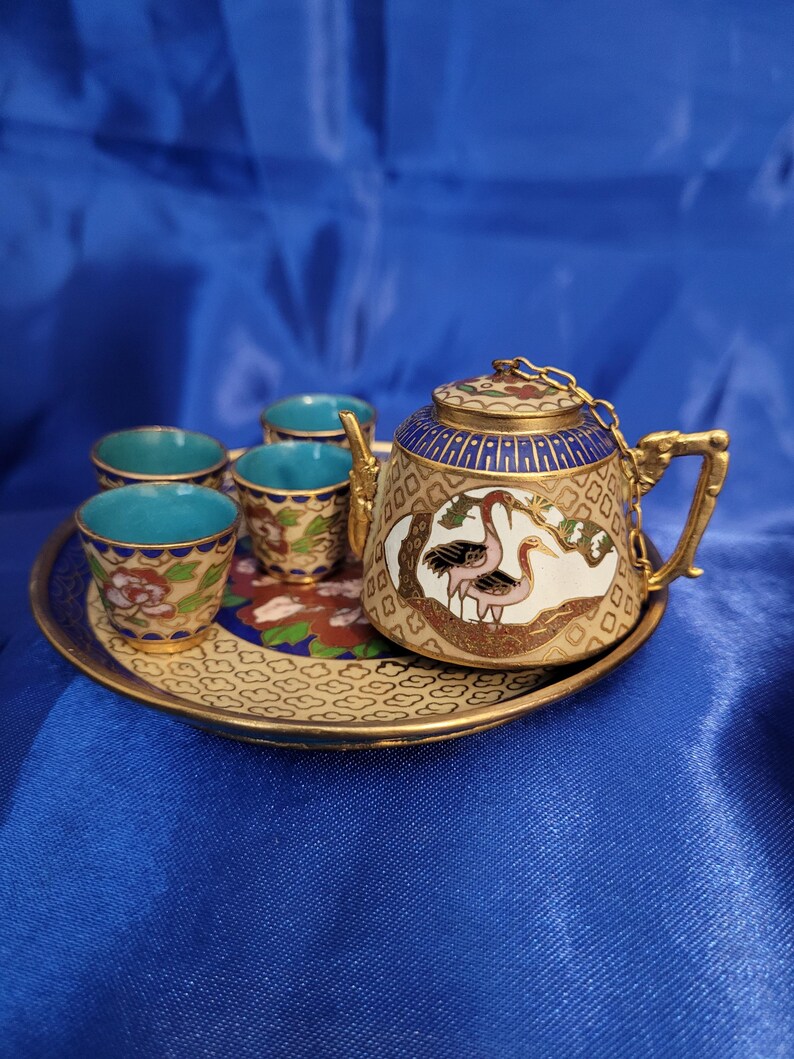 May include: Miniature tea set with a gold-colored tray, teapot, and four cups. The teapot and cups have a teal interior and are decorated with gold, blue, and red floral patterns. The teapot features a crane design.