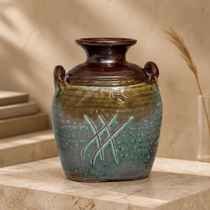 Puede incluir: Un jarrón de cerámica hecho a mano con la parte superior y asas de color marrón oscuro. El cuerpo del jarrón es una mezcla de tonos turquesa, marrón y verde, con un diseño grabado. El jarrón es una pieza decorativa.