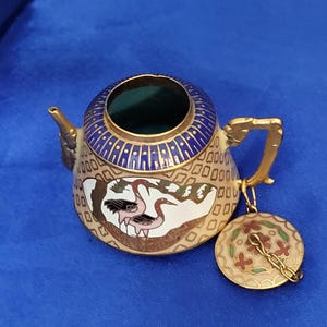 May include: A small, decorative teapot with a lid. The teapot is beige with a blue and gold rim and handle. It features a painted design of a crane and floral patterns. The lid is round and attached with a gold chain.