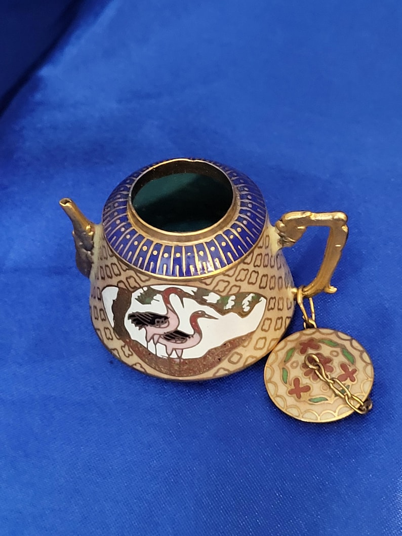 May include: A small, decorative teapot with a lid. The teapot is beige with a blue and gold rim and handle. It features a painted design of a crane and floral patterns. The lid is round and attached with a gold chain.