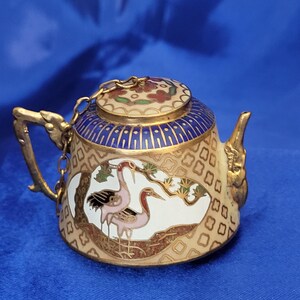 May include: Miniature decorative teapot with a gold handle and spout. The teapot features a cream-colored body with a blue and gold lid. The body is decorated with a crane design in pink, black, and gold. A gold chain connects the lid to the handle.
