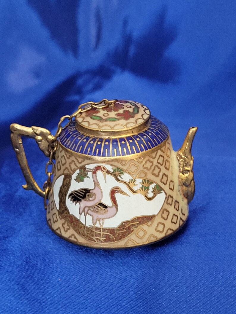 May include: Miniature decorative teapot with a gold handle and spout. The teapot features a cream-colored body with a blue and gold lid. The body is decorated with a crane design in pink, black, and gold. A gold chain connects the lid to the handle.
