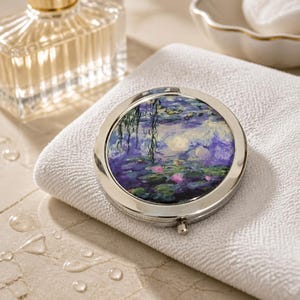 May include: A round, silver compact mirror featuring a painting of water lilies in shades of purple, blue, and green. The mirror is resting on a white towel, with a perfume bottle and a small bowl in the background. The compact has a small button to open it.