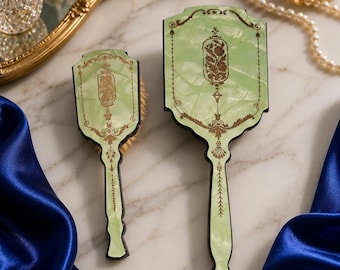 May include: A vintage-style vanity set featuring a brush and a hand mirror. The set has a light green, marbled finish with ornate gold-colored detailing. The brush has soft bristles, and the mirror has a decorative floral design.