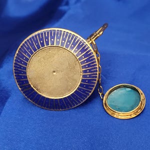 May include: A gold-toned, circular pendant with a blue enamel border and a central, unadorned area. A small, oval-shaped pendant with a turquoise stone is attached by a delicate chain. The background is a solid blue.