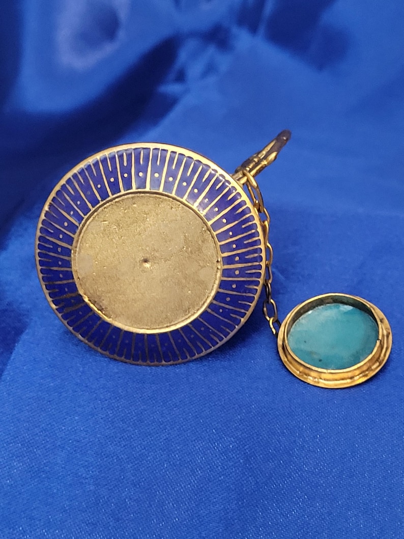 May include: A gold-toned, circular pendant with a blue enamel border and a central, unadorned area. A small, oval-shaped pendant with a turquoise stone is attached by a delicate chain. The background is a solid blue.