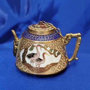 May include: A small, ornate teapot with a gold finish, featuring a blue and white patterned lid and body. The teapot has a chain connecting the lid to the handle. The body of the teapot has a painted scene of two pink cranes.