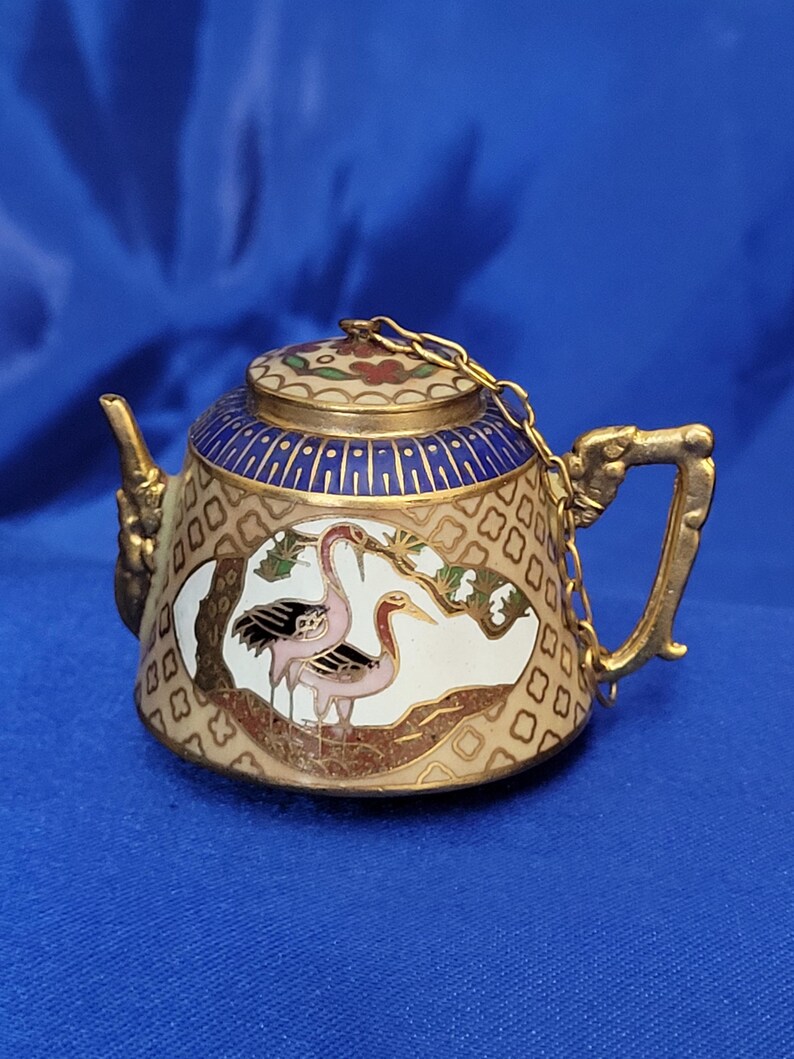 May include: A small, ornate teapot with a gold finish, featuring a blue and white patterned lid and body. The teapot has a chain connecting the lid to the handle. The body of the teapot has a painted scene of two pink cranes.