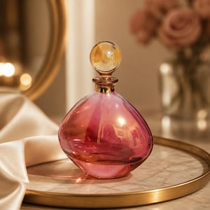 May include: A decorative glass perfume bottle with a pink and orange gradient. The bottle has a round, bulbous shape with a gold-colored rim and a spherical stopper. The bottle sits on a gold-rimmed tray.