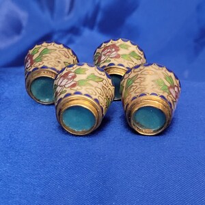 May include: Four decorative thimbles with floral designs. The thimbles have a cream-colored base with red and green floral patterns, gold trim, and a blue interior. They are arranged on a blue surface.