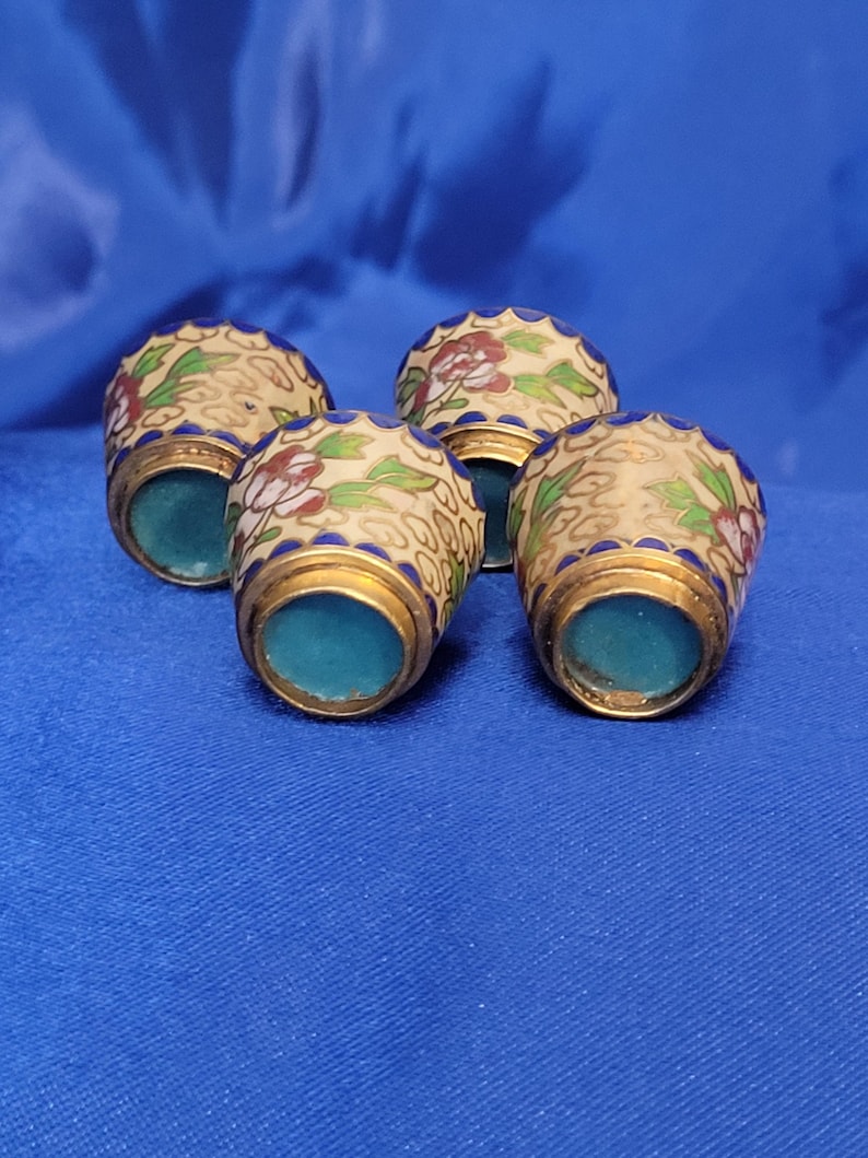 May include: Four decorative thimbles with floral designs. The thimbles have a cream-colored base with red and green floral patterns, gold trim, and a blue interior. They are arranged on a blue surface.